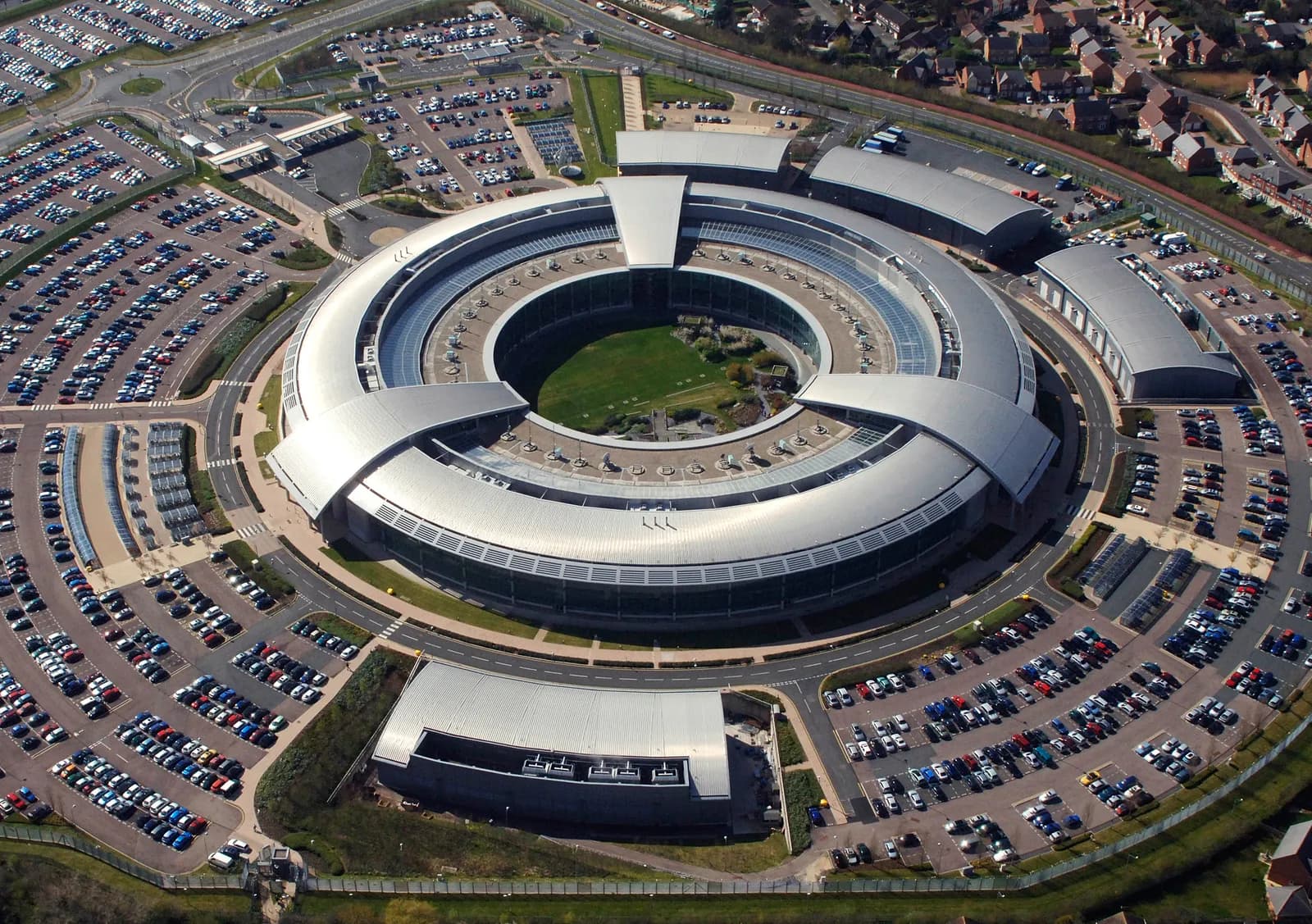 An aerial view shows the circular, modern building of GCHQ surrounded by several parking lots filled with cars.