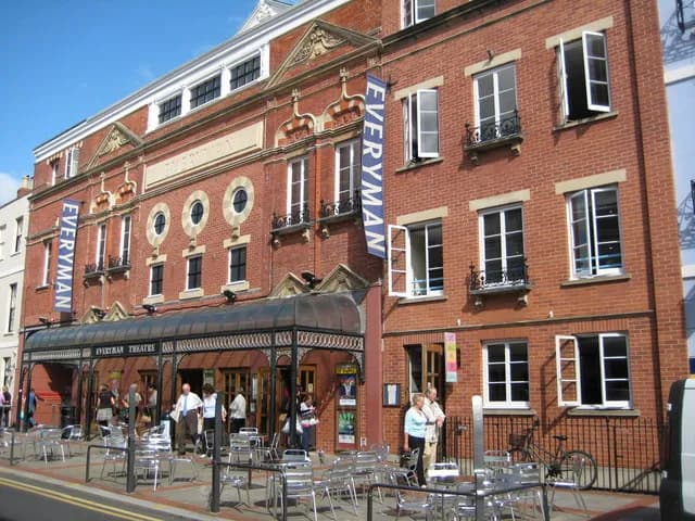 A red brick building with the name "Everyman Theatre" visible on banners and signage, featuring multiple windows, a shaded entrance area, and people gathered outside.