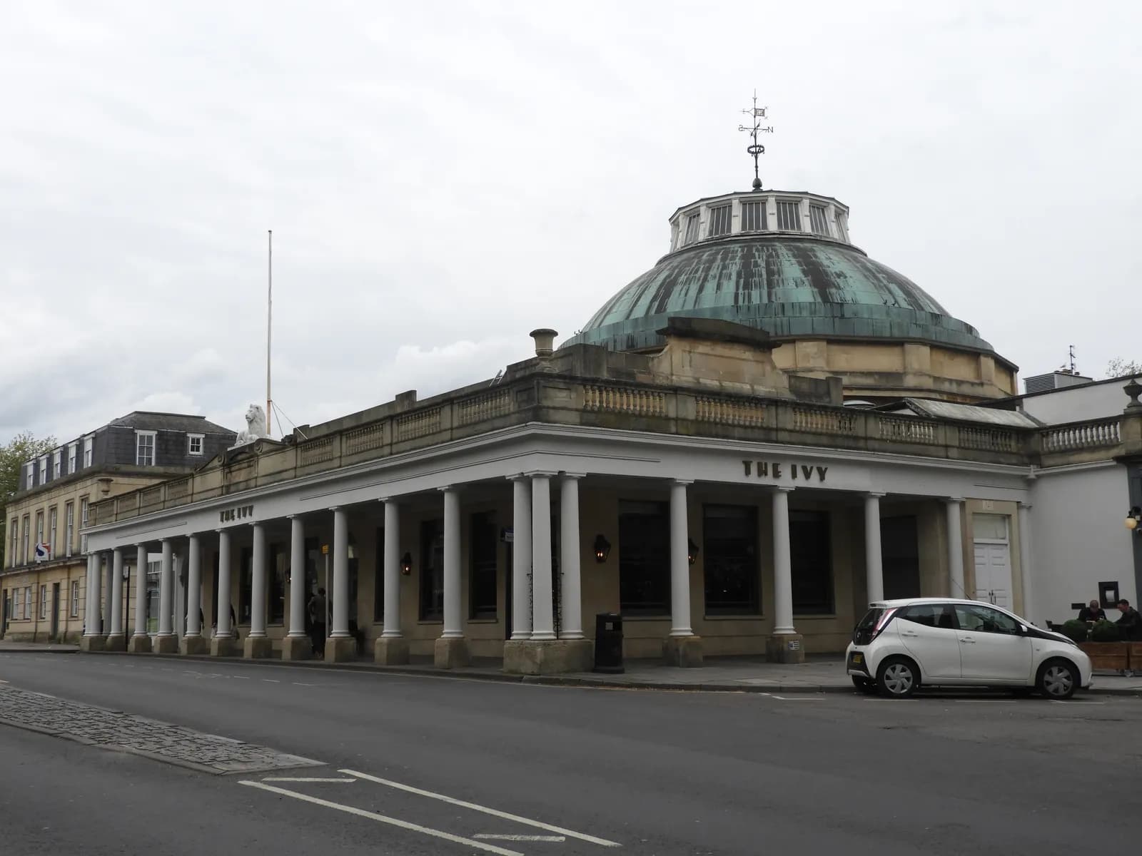 The Ivy restaurant and bar is housed in a grand historic building with a domed roof and a colonnade of pillars along a street with a white car parked in front.
