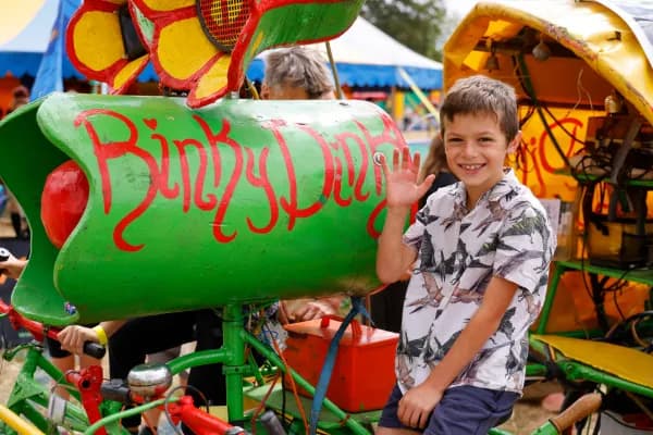 A smiling boy in a patterned shirt waves while sitting next to a green-painted vehicle labeled "Binky's Dirty" with a red object in its side.