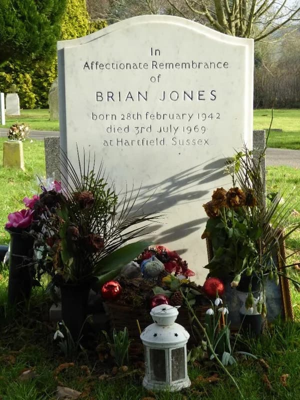 A white headstone for Brian Jones in a grassy cemetery, adorned with flowers, ornaments, and a small white lantern at its base.